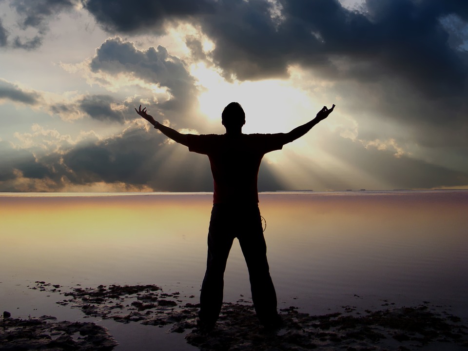 Silhouette of a person on a beach with arms raised as sunbeams break through cloudy skies.