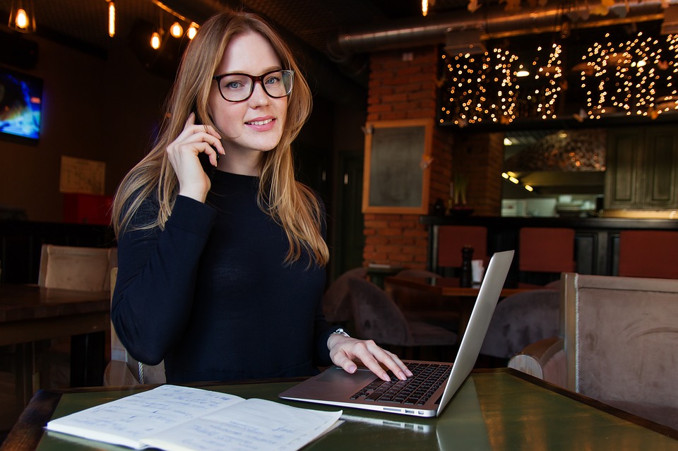 Woman wearing glasses sits at a café table, talking on a phone while typing on a laptop with a notebook open nearby.