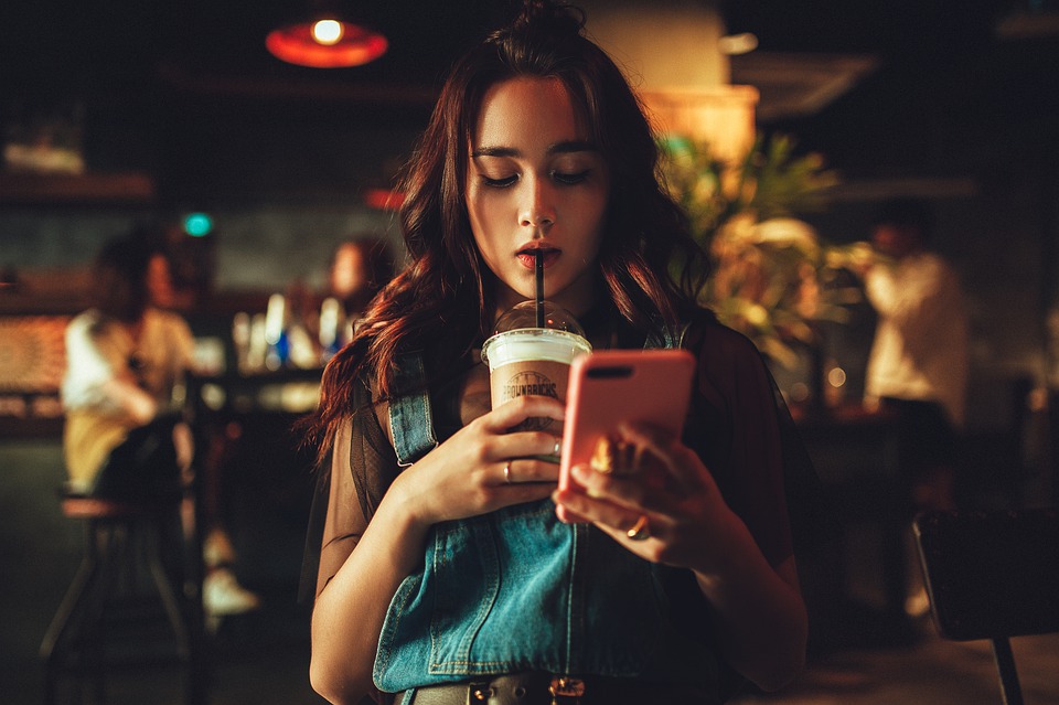 Woman in a cafe sips a iced drink through a straw while looking at her pink smartphone.